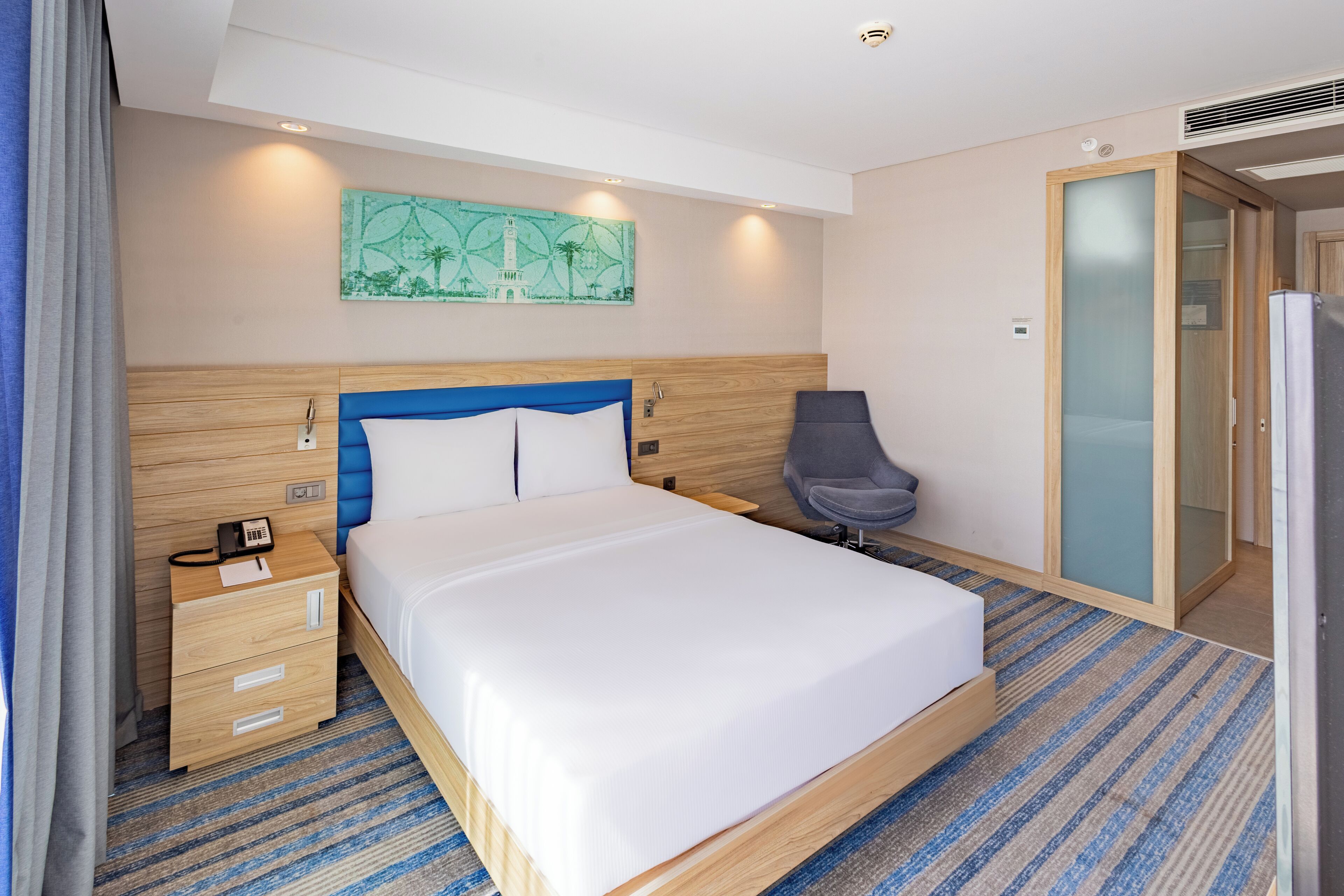 A hotel room with a large bed, a bedside table with a phone, a chair, and a wardrobe.
