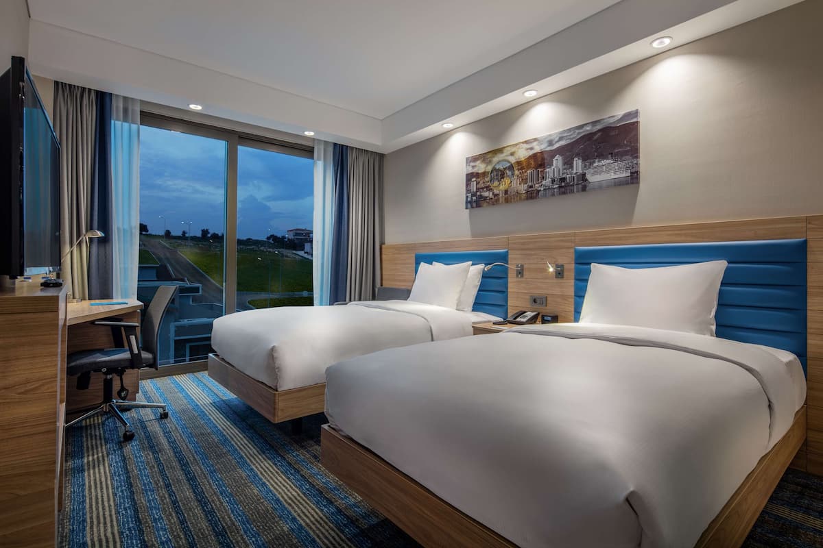Hotelkamer met twee bedden, een bureau en een groot raam met uitzicht op een weg en het landschap.