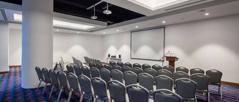 Une salle de conférence avec des rangées de chaises, un pupitre, un écran de projection et un sol recouvert de moquette.