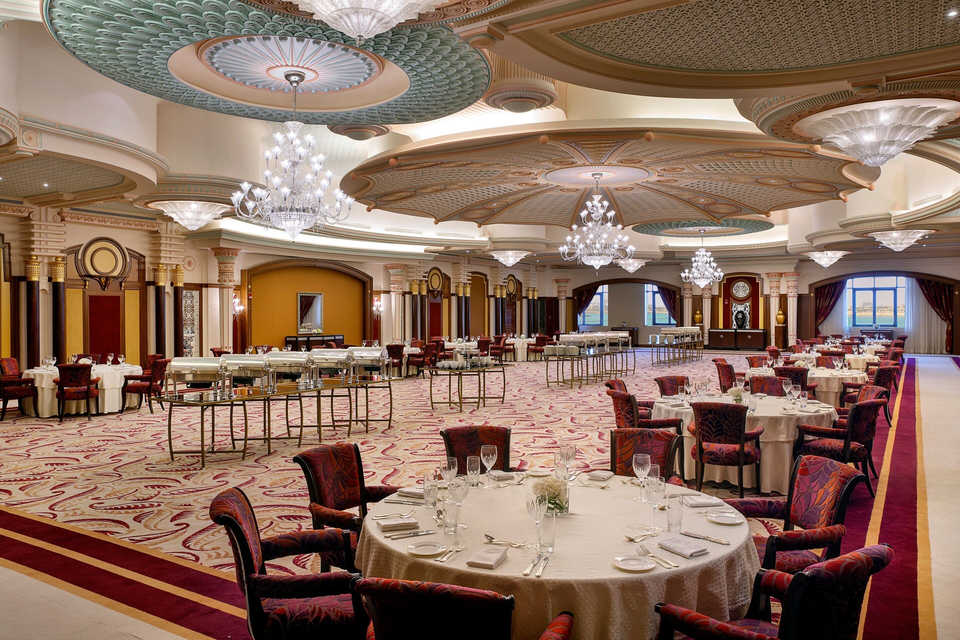 A spacious banquet hall with round tables set for dining, ornate chandeliers, and a grand ceiling design.