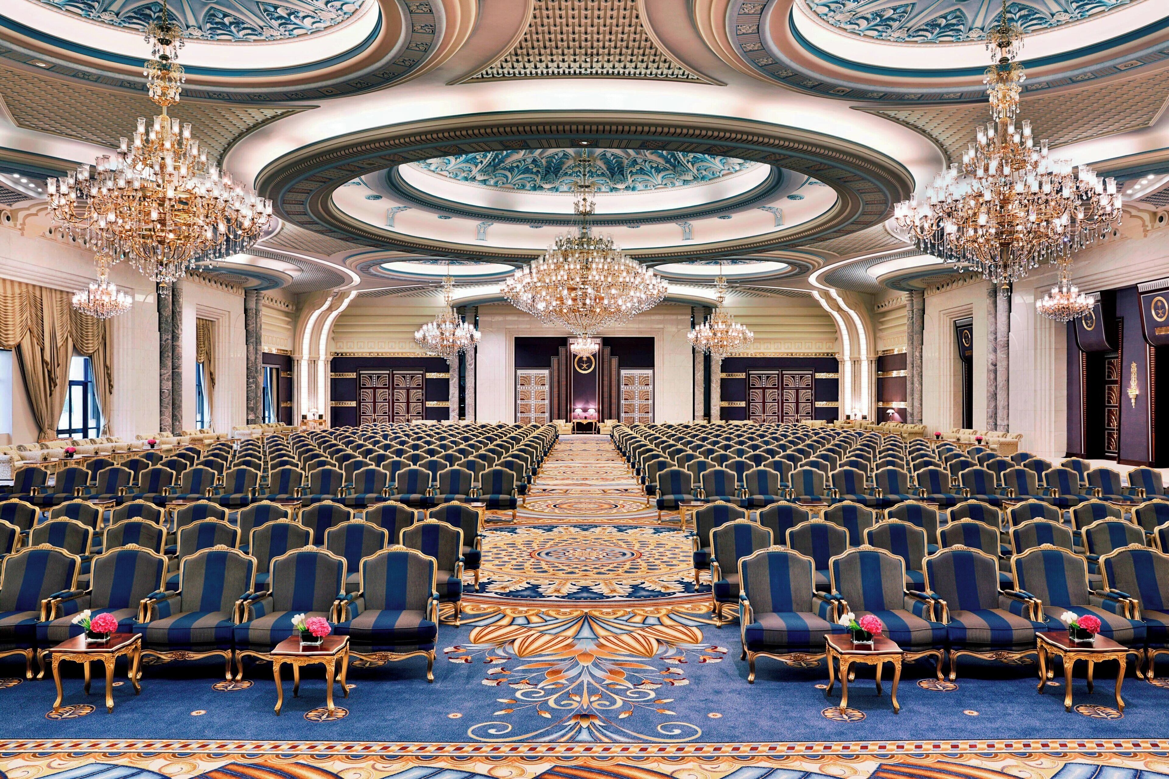 Un gran salón con hileras de sillas tapizadas en azul, candelabros ornamentados y una alfombra detallada.