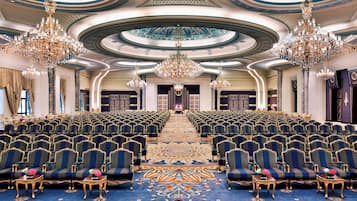 Un gran salón con hileras de sillas tapizadas en azul, candelabros ornamentados y una alfombra detallada.