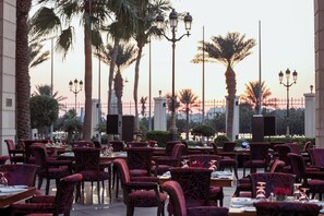 An outdoor dining area with red upholstered chairs and tables, palm trees, and street lamps.
