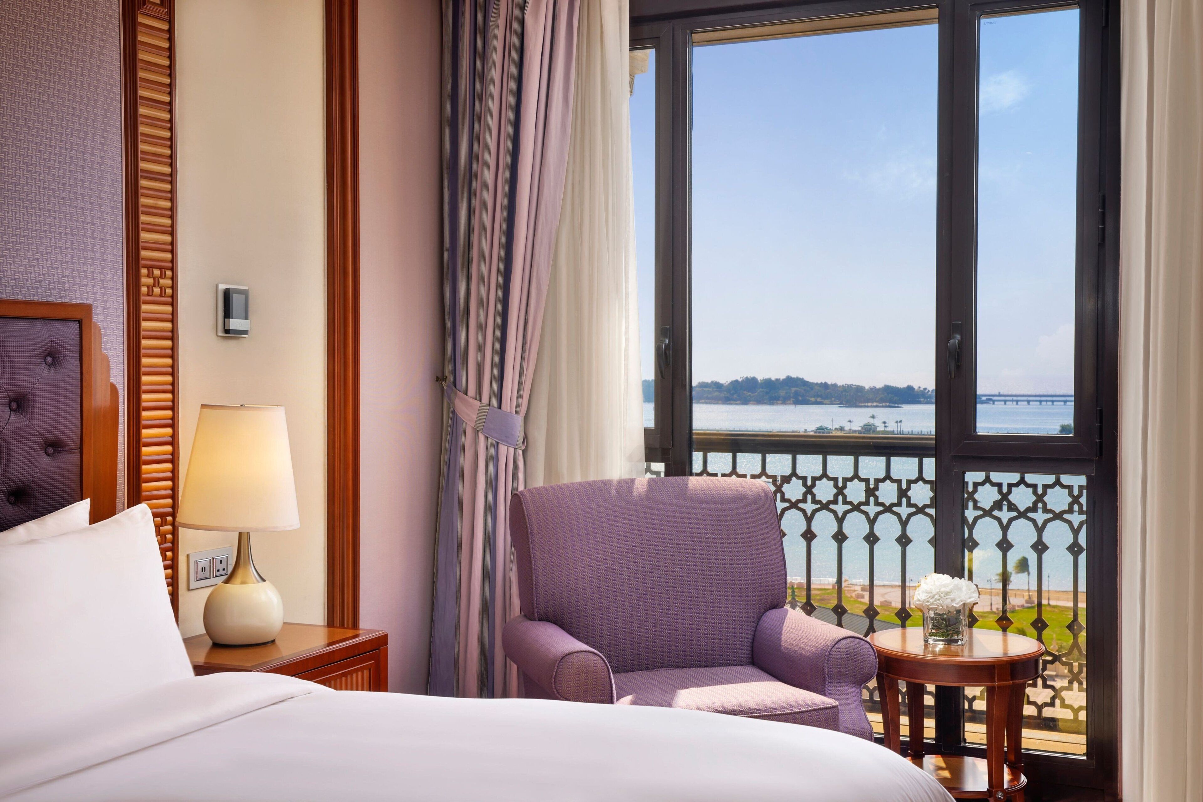 A hotel room with a bed, a chair, a small table with a vase, and a view of the sea through a balcony.