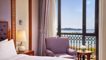 A hotel room with a bed, a chair, a small table with a vase, and a view of the sea through a balcony.