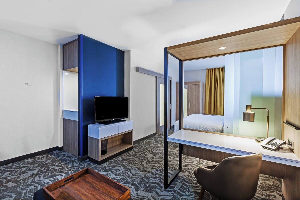 A hotel room with a desk, a television, a bed, a mirror, and a small table.