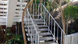 A staircase with white railings leading to an upper level, surrounded by tropical plants and a thatched roof structure. - La Mer Guesthouse (Gqeberha (Port Elizabeth))