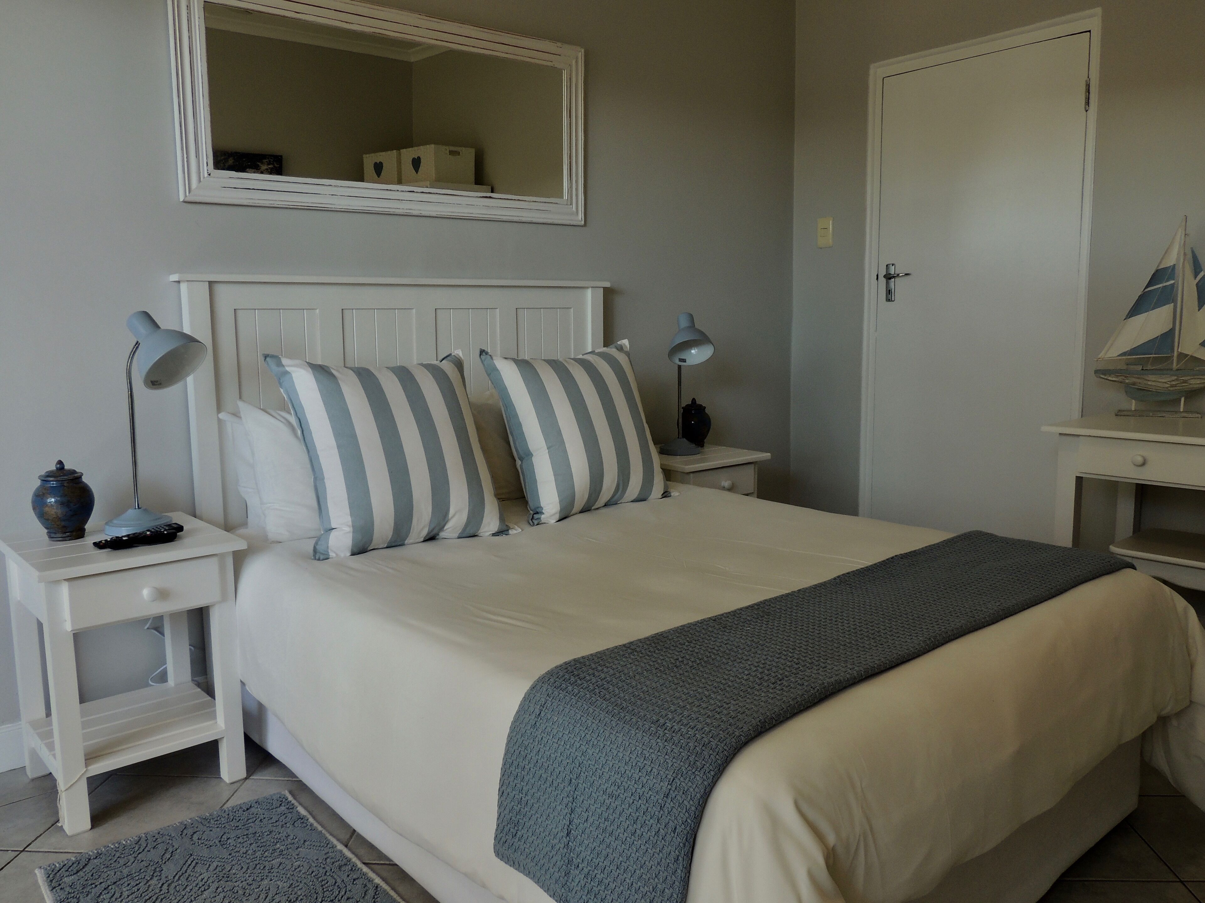 A neatly arranged bedroom with a bed, two bedside tables, a nightstand, a mirror, and a small decorative sailboat.