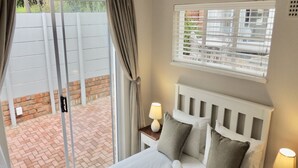 A neatly made bed with pillows and a blanket, a bedside table with a lamp, a window with blinds, and a view of a brick patio outside. - La Mer Guesthouse (Gqeberha (Port Elizabeth))