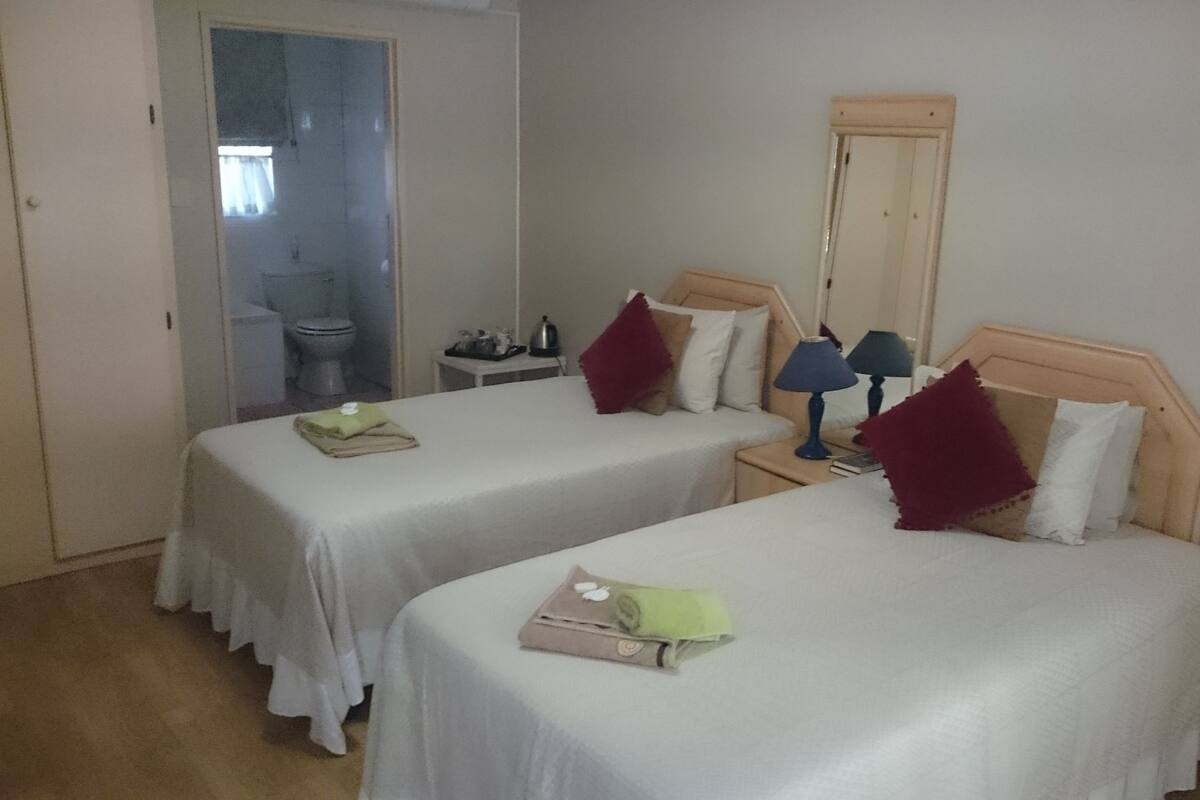 A hotel room with two single beds, a small table with a lamp, a mirror, and a bathroom visible through an open door.