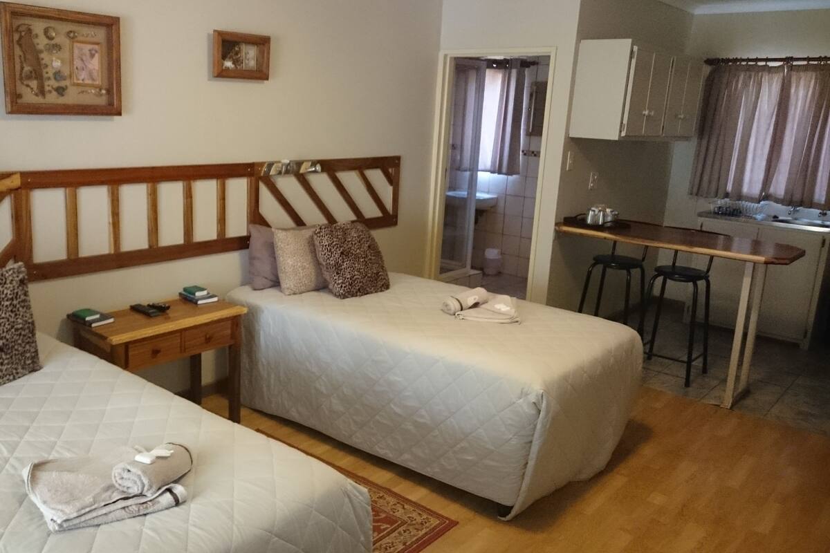A hotel room with two beds, a desk, a chair, a small kitchenette, and a bathroom visible through an open door.