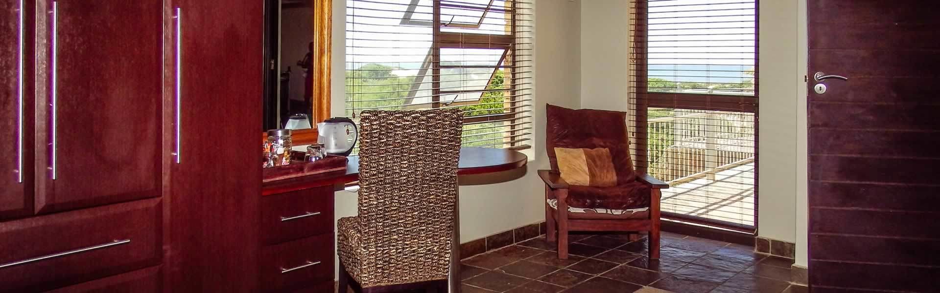 A room with a wooden cabinet, a woven chair, a cushioned chair, and a view through a window with blinds.