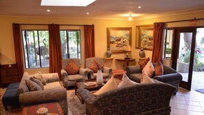 Amplia sala de estar con varios sofás y sillones, una mesa de centro y una estantería. Grandes ventanales con cortinas dejan entrar luz natural y ofrecen vistas al exterior.