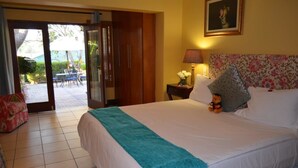 Una habitación de hotel con una cama grande, una mesita de noche con lámpara, un cabecero floral y un ventanal corredero que da a un espacio exterior con mesa y sillas.