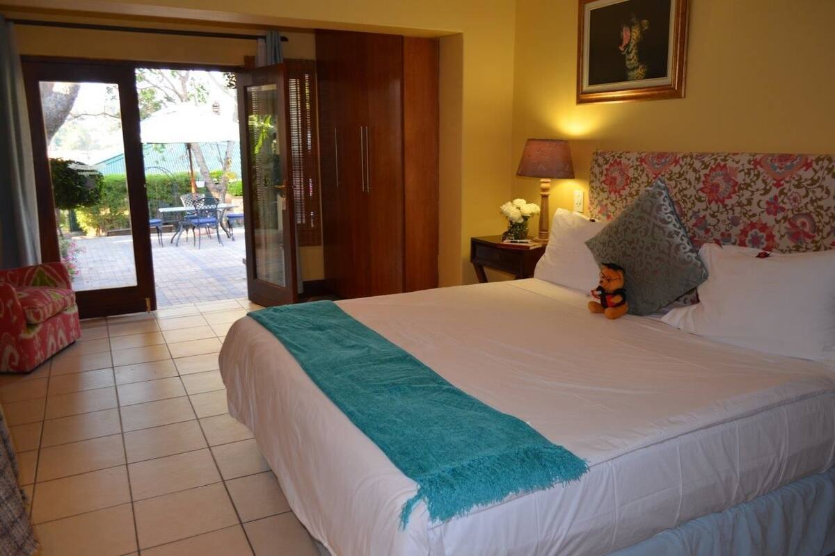 A hotel room with a large bed, a nightstand with a lamp, a floral headboard, and a sliding glass door leading to an outdoor area with a table and chairs.