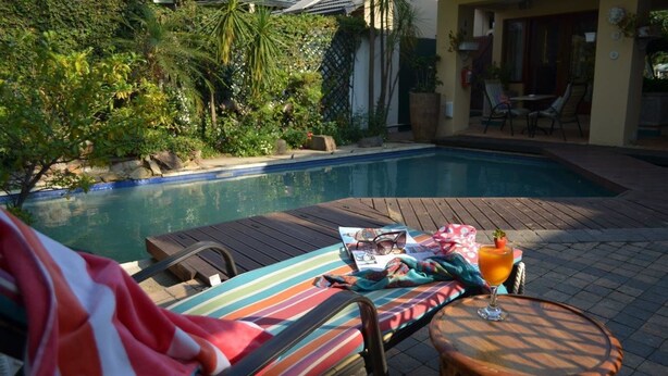 Zona junto a la piscina con una tumbona, una mesita con una bebida y un porche cubierto.
