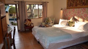 Un dormitorio con una cama grande, una mesa de comedor y una silla. Hay una ventana con cortinas, un cuadro en la pared y un árbol pequeño afuera.