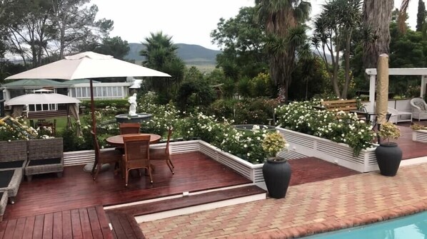 A poolside area with a wooden deck, outdoor furniture, a white umbrella, and a garden with white flowers. - Tri-Angle B&B (Nxuba)