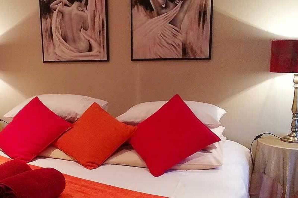 Una camera d'albergo con un letto, cuscini rossi e bianchi, due lampade sui comodini, un tavolino con una lampada e una TV a muro.