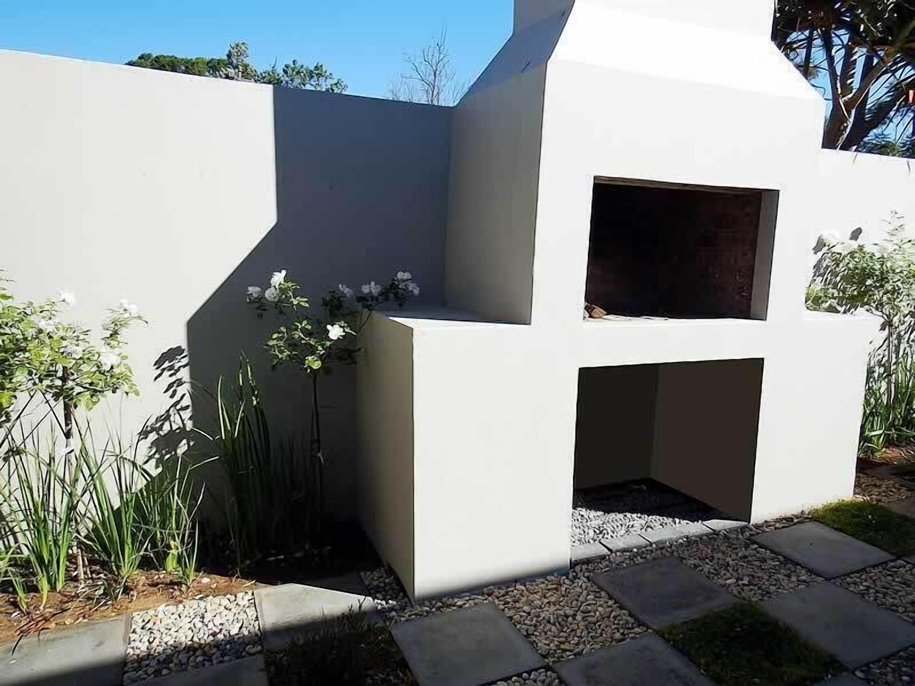 Un espace cuisine extérieur moderne, doté d’une structure blanche, d’un barbecue encastré et entouré de plantes.