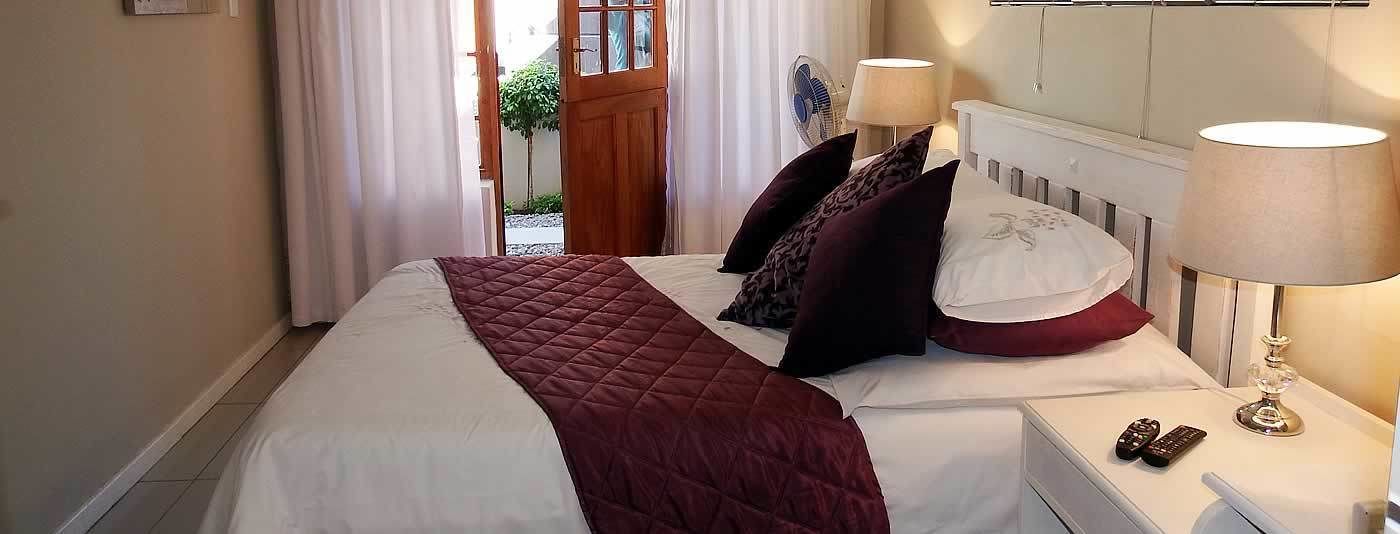 A neatly made bed with a white and maroon bedspread, two pillows, and a bedside table with a lamp and remote controls.