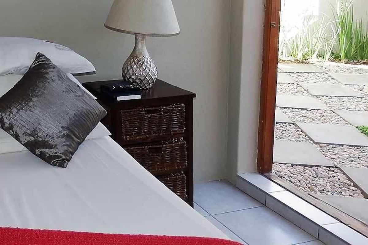 Un letto rifatto con cura, lenzuola bianche e dettagli rossi, un comodino in legno scuro accanto e una lampada sopra il comodino.
