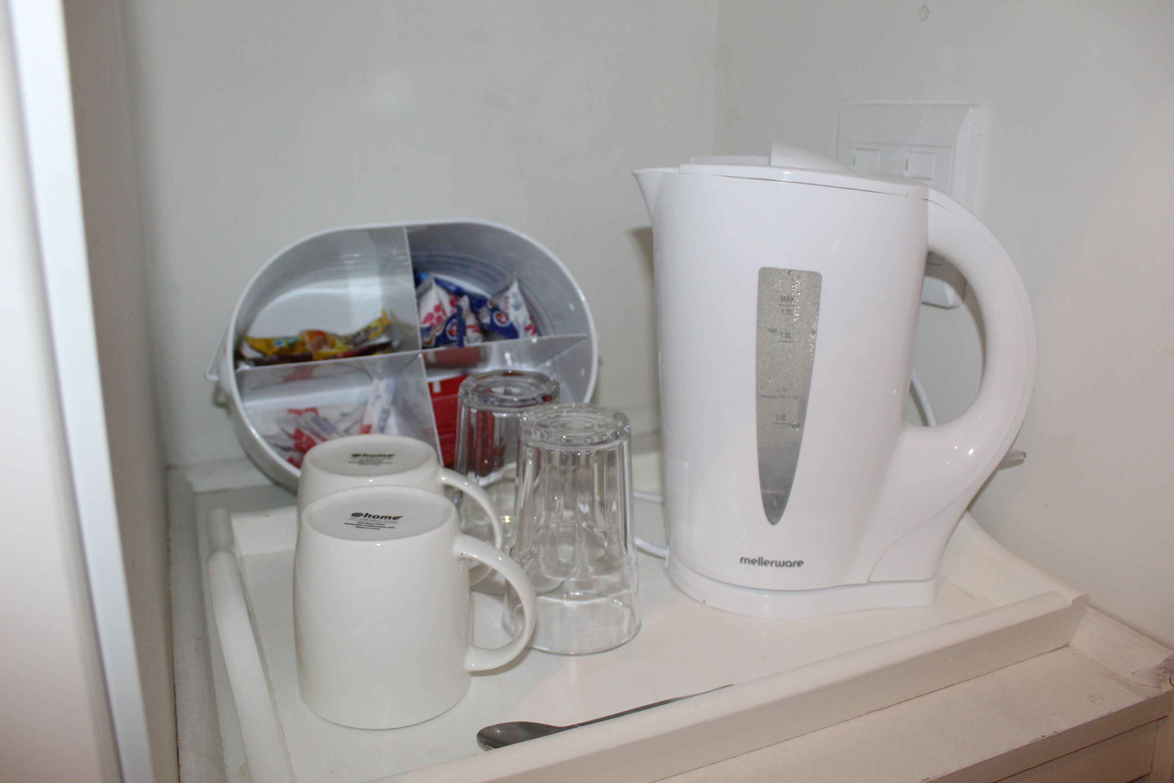 Una kitchenette con un hervidor blanco, una jarra de vidrio, una taza y un recipiente con snacks.