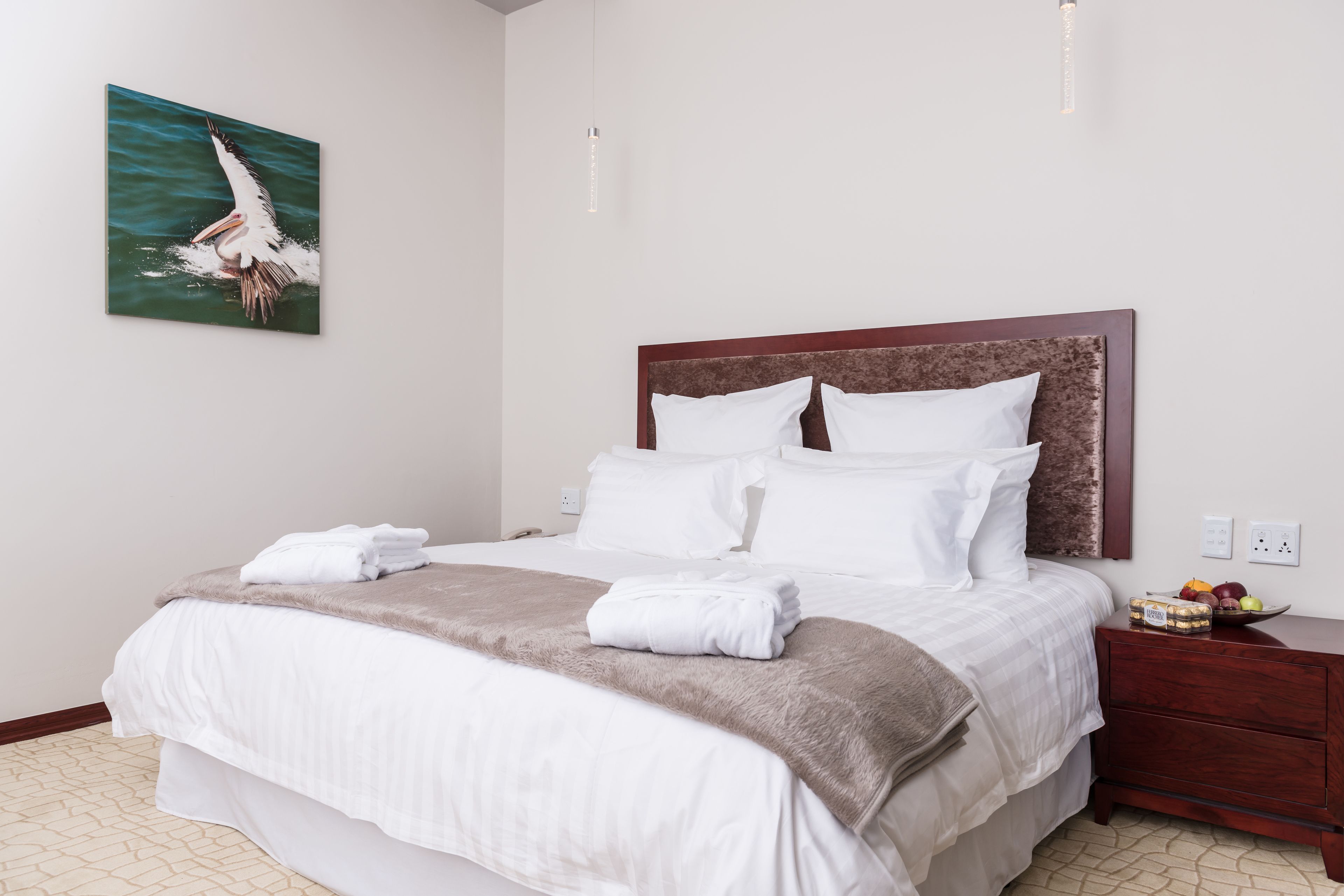 Een net opgemaakt bed met witte lakens en een bruine plaid, een houten nachtkastje met een schaal fruit, en een schilderij van een vogel aan de muur.