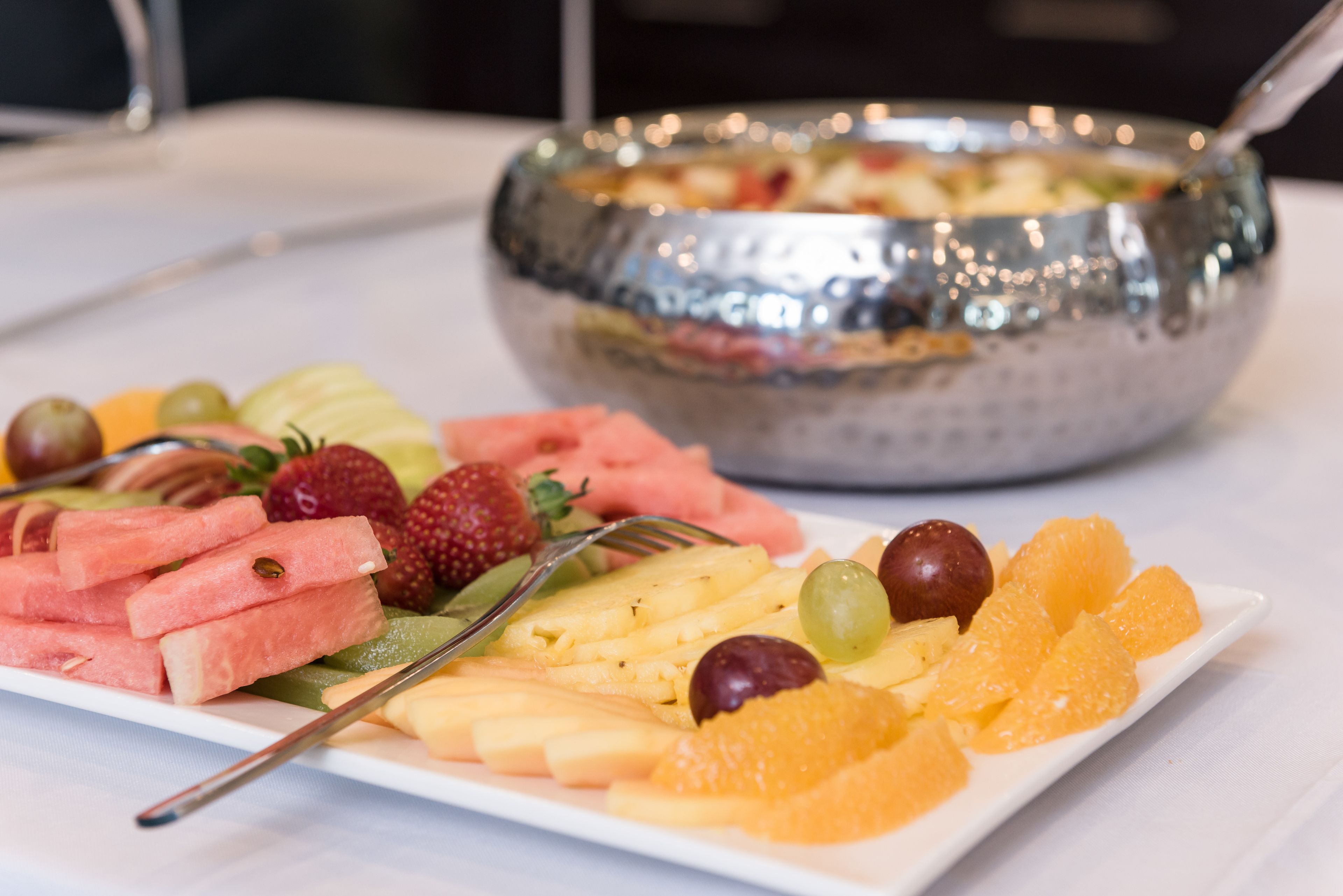 A white plate with various fruits and a fork, and a metal bowl with a spoon in the background.
