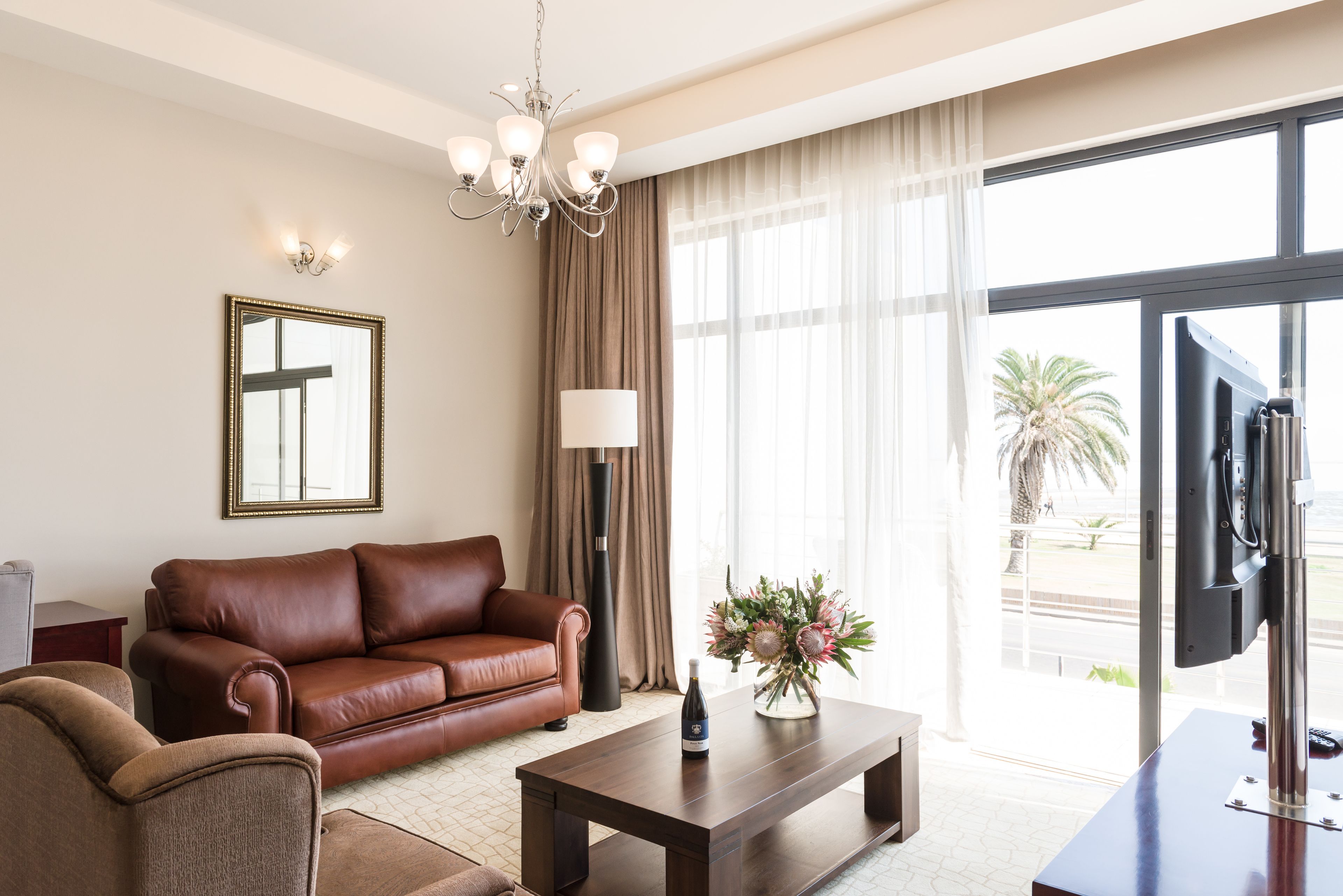 Una sala de estar moderna con un sofá de cuero, una mesa de centro con una botella, un jarrón con flores, un espejo grande y un televisor sobre un mueble.