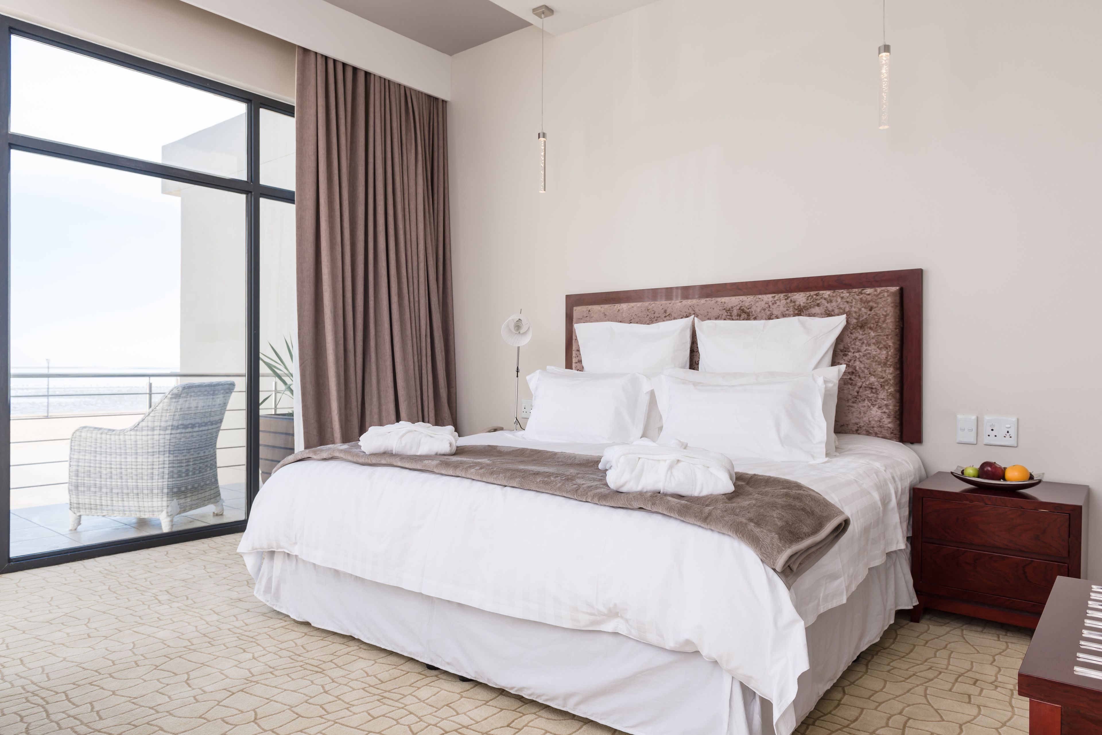 A hotel room with a large bed, a nightstand with a fruit bowl, a chair by the window, and a view of the outside.