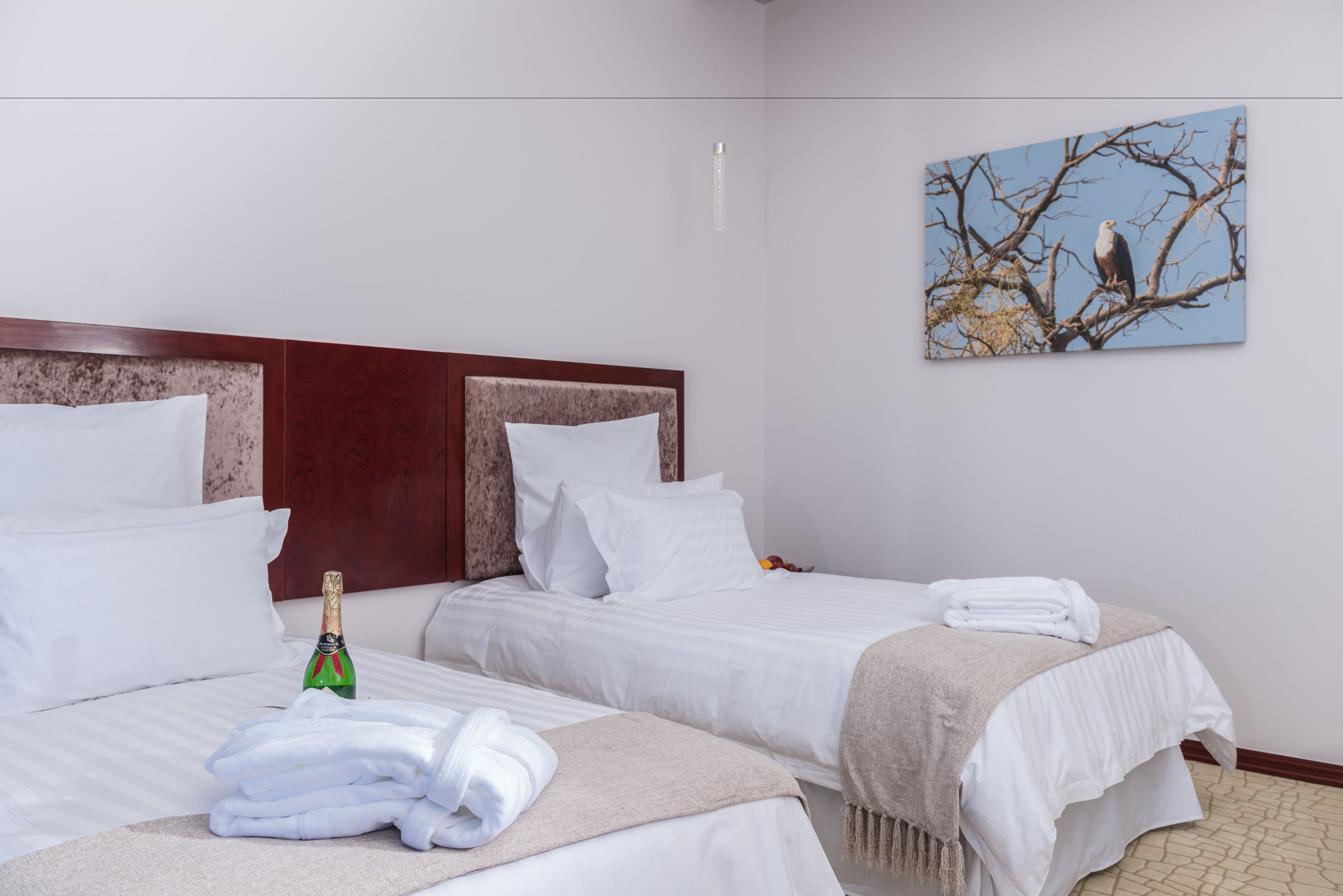 Une chambre d’hôtel avec deux lits, du linge de lit blanc, une bouteille de champagne et un tableau représentant un oiseau perché sur une branche.