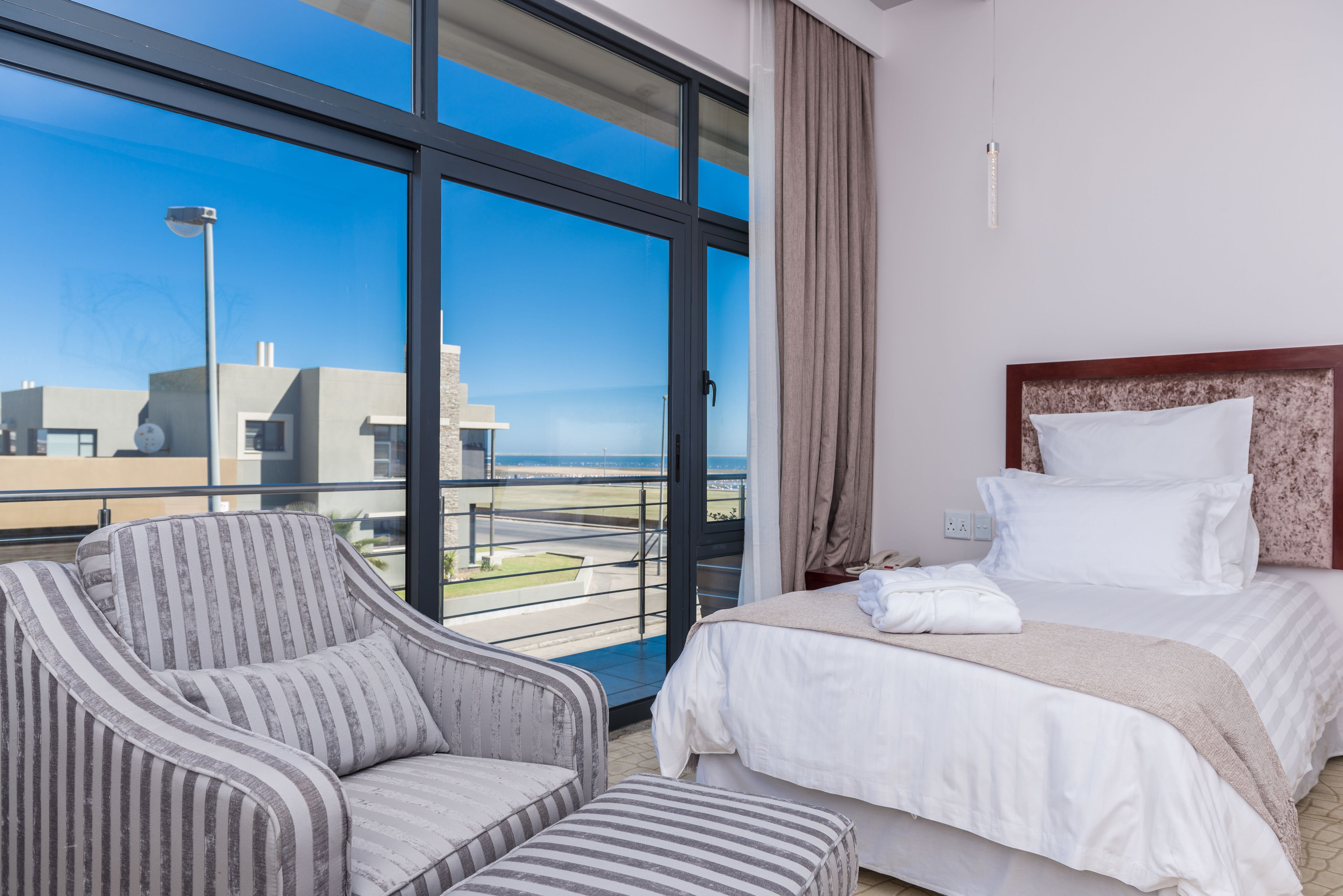 Una habitación de hotel con un ventanal grande, una cama con sábanas blancas, un sillón a rayas y vistas a la ciudad.