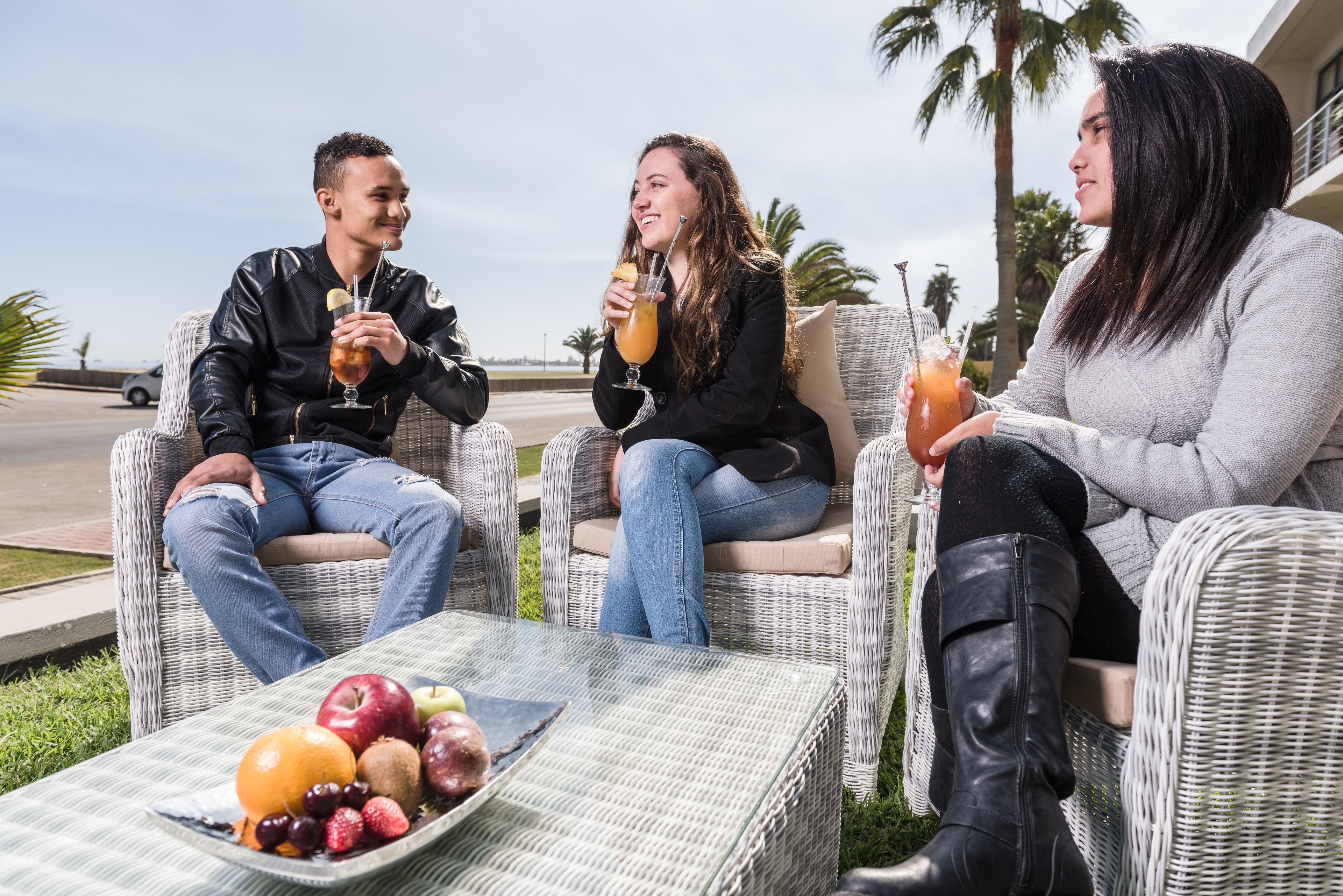 Três pessoas sentadas em cadeiras de vime ao ar livre, com uma mesa de frutas e bebidas à frente.