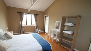 Een slaapkamer met een bed, een bureau, een stoel, een ventilator en een plank met handdoeken.
