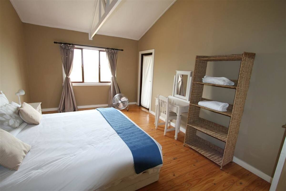 Een slaapkamer met een bed, een bureau, een stoel, een ventilator en een plank met handdoeken.