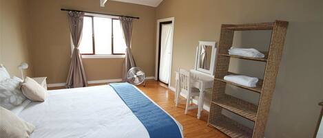 A bedroom with a bed, a desk, a chair, a fan, and a shelf with towels.