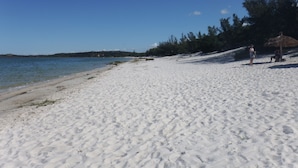 Een zandstrand met helderblauw water, enkele bomen en een rieten parasol.
