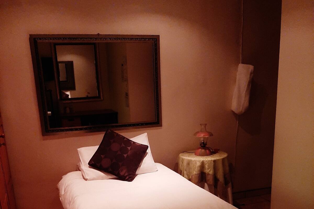 A single bed with a pillow, a bedside table with a lamp, and a bench in a dimly lit room with tiled flooring.