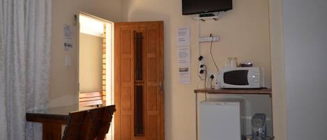 A compact room with a wooden door, a small white refrigerator, a microwave on a shelf, a television on the wall, and a dining table with chairs.