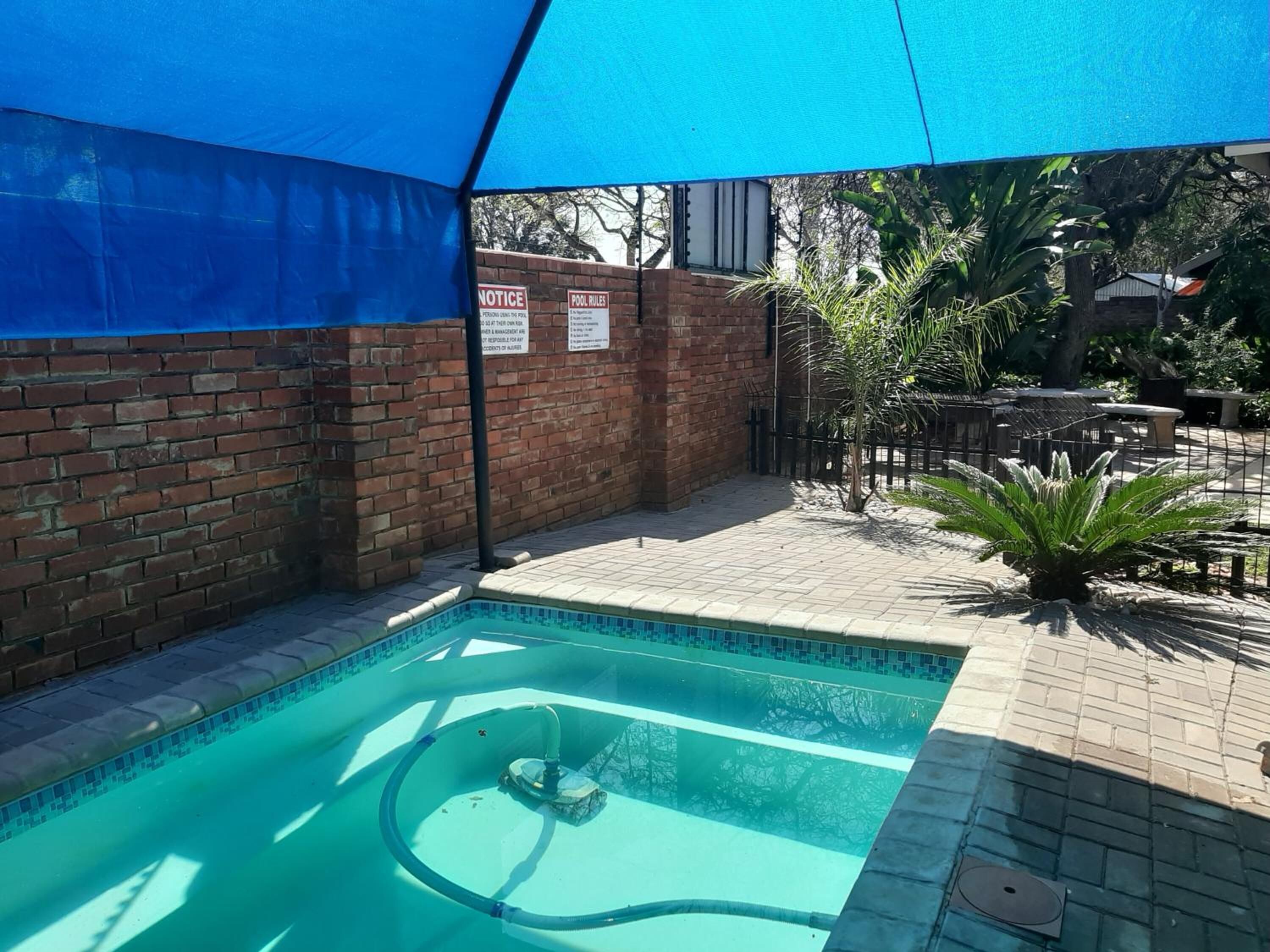En swimmingpool med ett blått solskydd ovanför, omgiven av en mur av tegelsten och en uteplats med kakel.