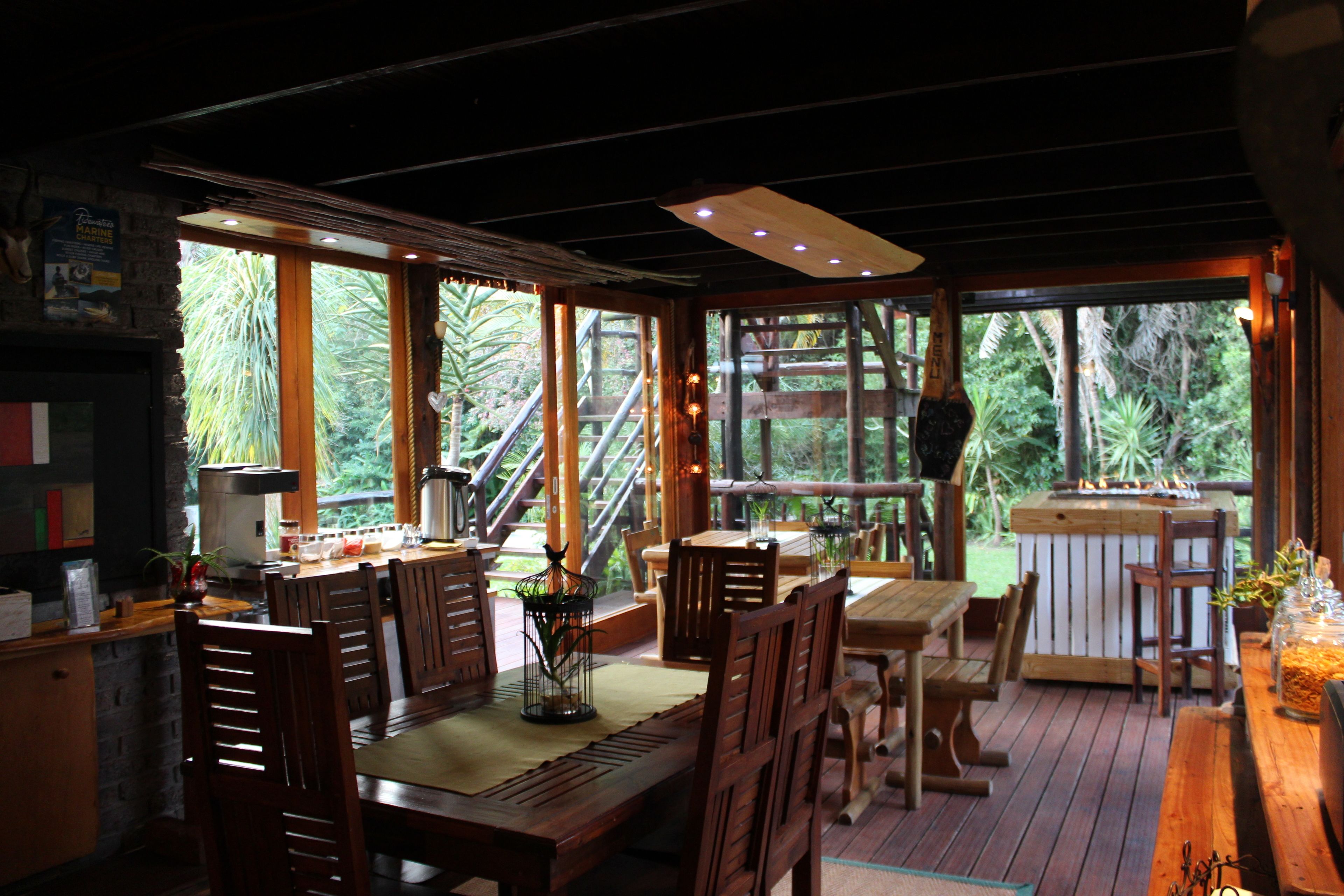 A wooden deck with dining tables and chairs, large windows with a view of greenery, and a wooden staircase leading outside.