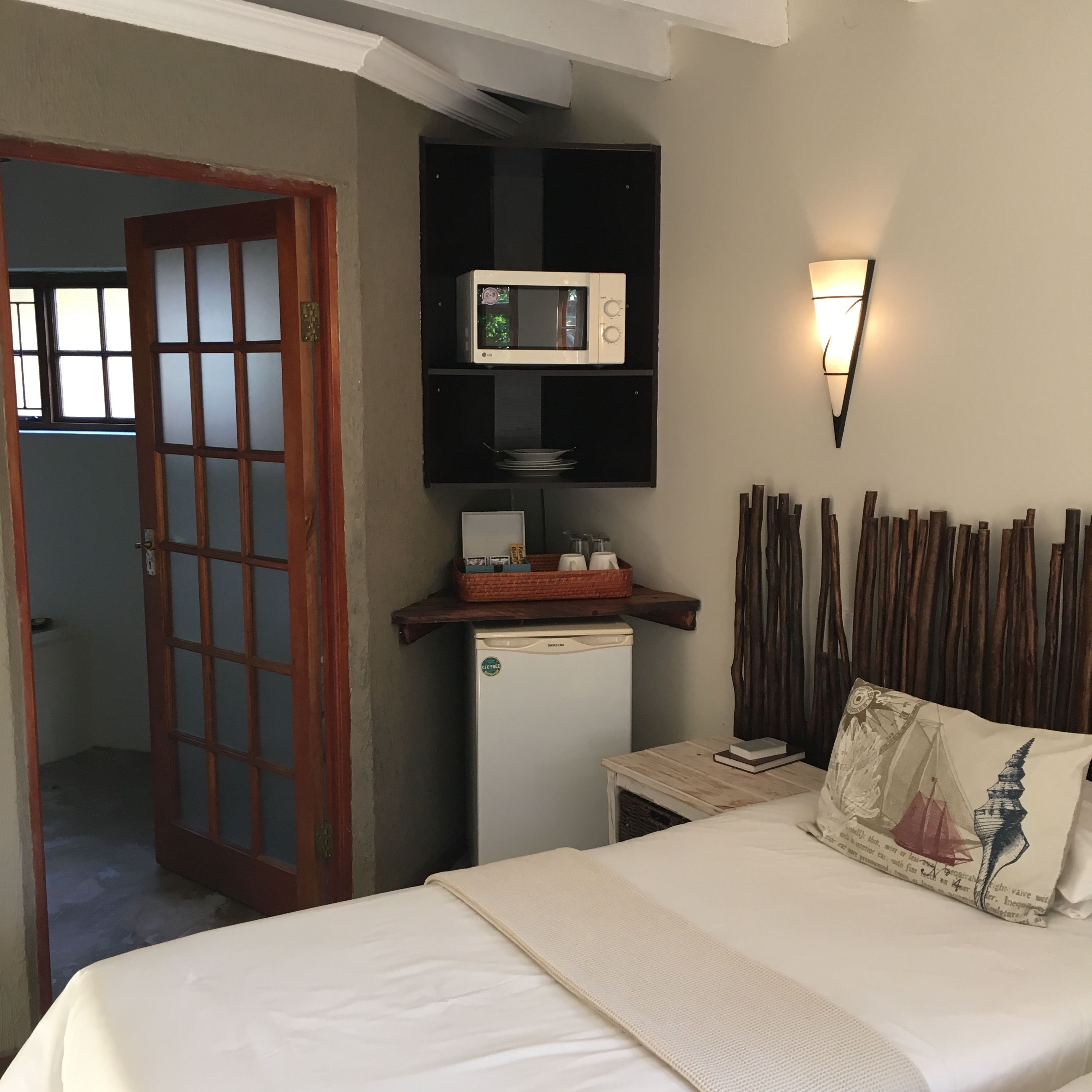 Un dormitorio con una cama, un refrigerador pequeño, un microondas, un estante en la pared con vajilla y un cojín decorativo.