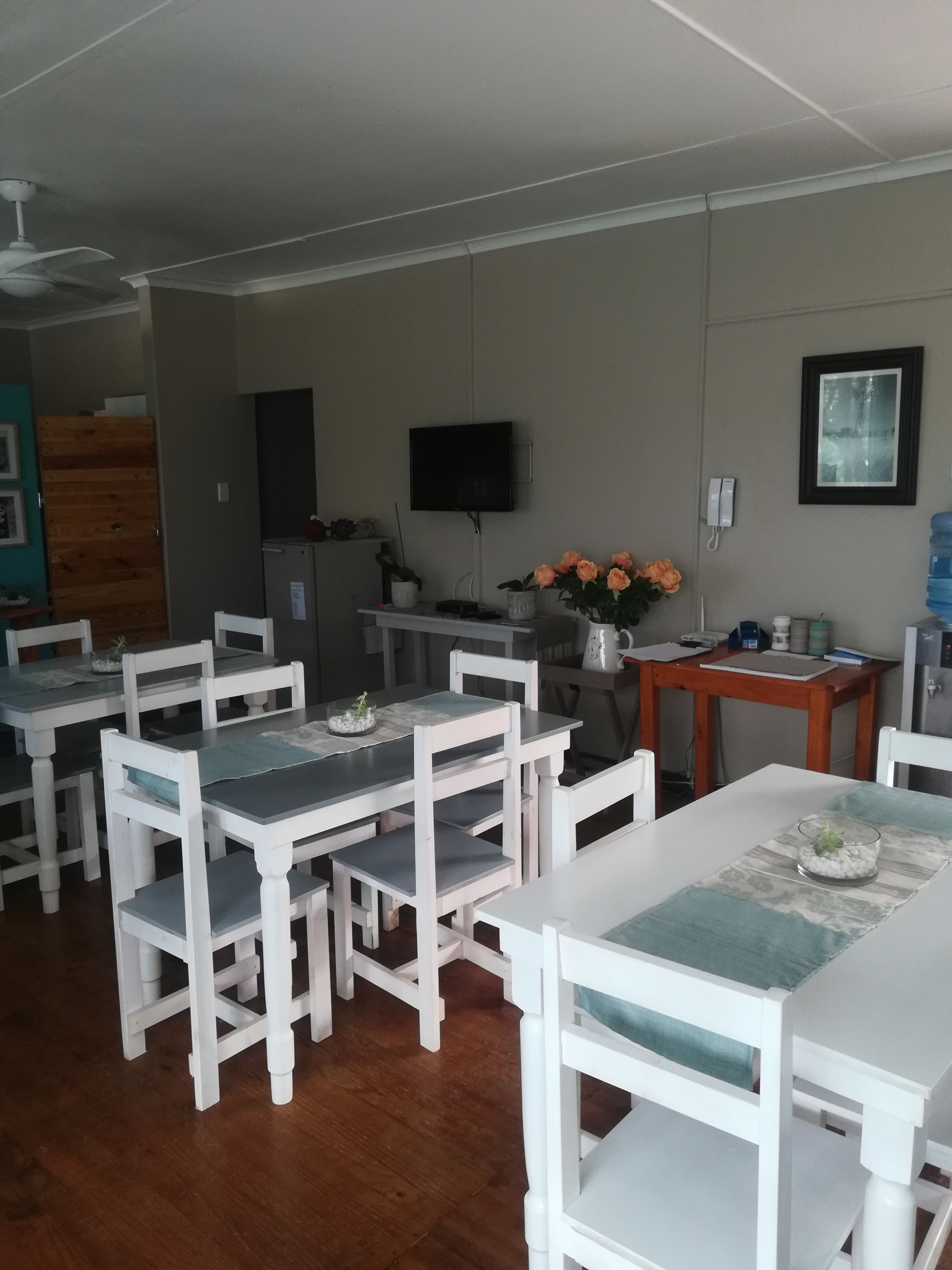 A dining area with white tables and chairs, a television mounted on the wall, and a water dispenser.