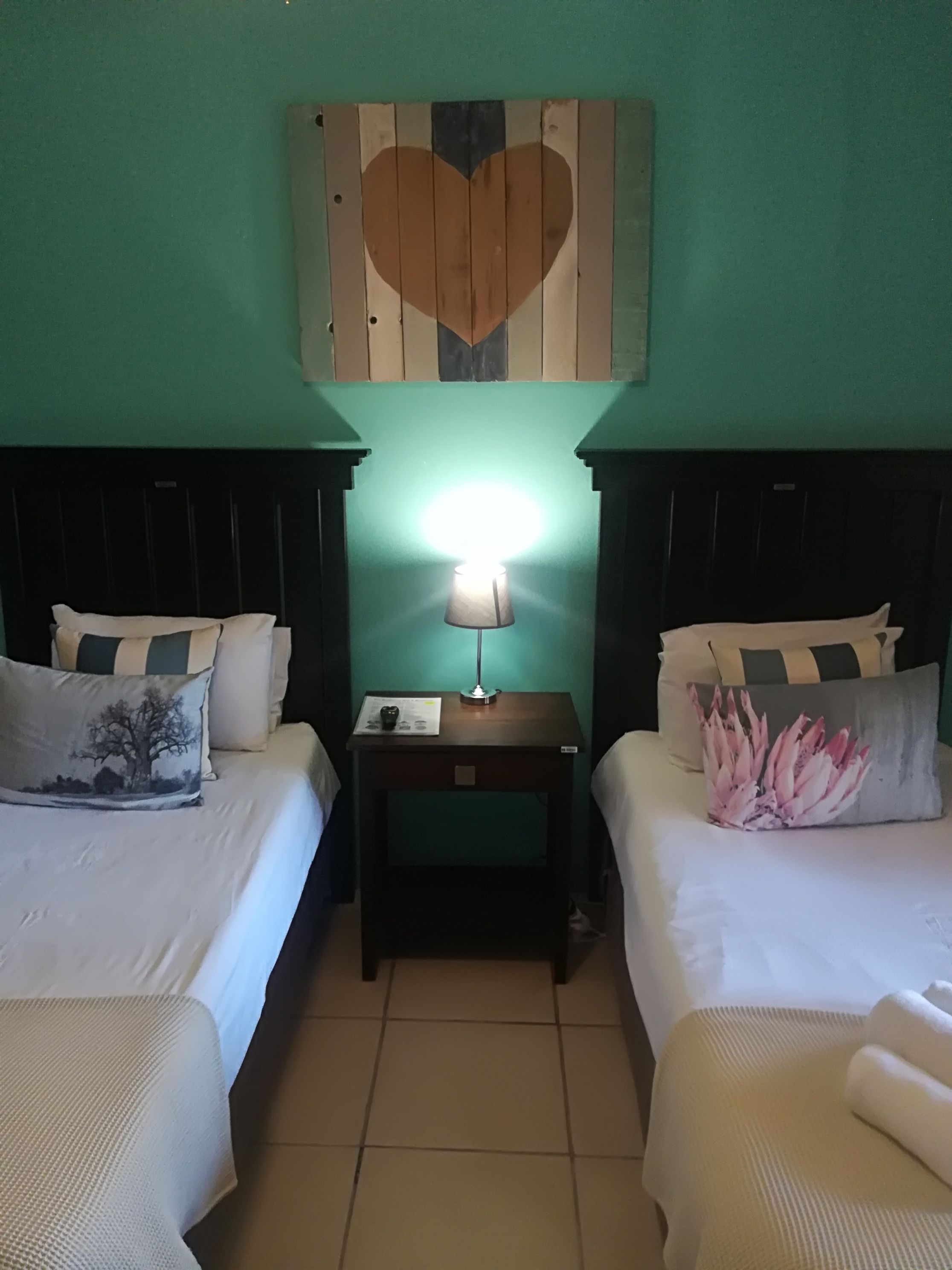 Una habitación con dos camas, una mesita de noche con una lámpara y un adorno de corazón en la pared.