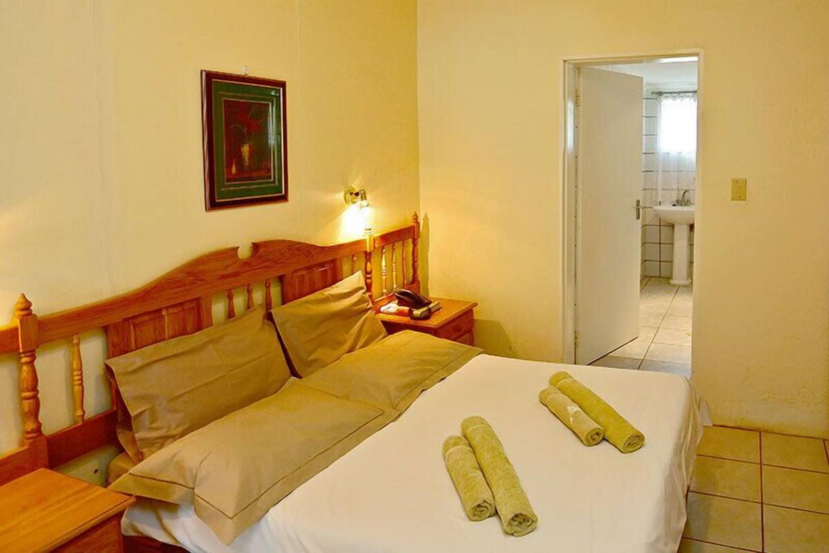 A hotel room with a wooden bed, a nightstand, a framed picture on the wall, and a bathroom visible through an open door.
