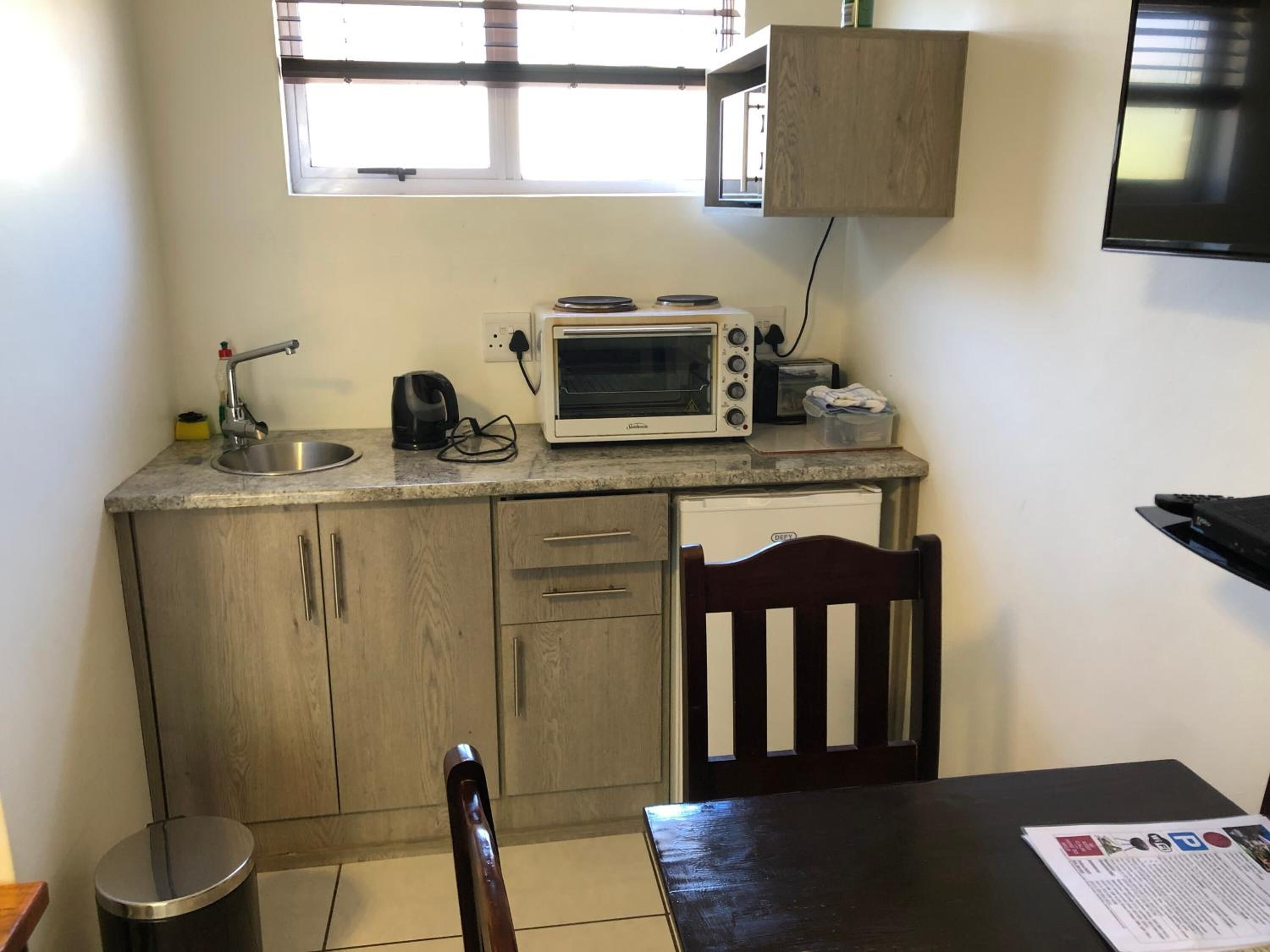 Una cocina compacta con microondas integrado, hervidor y lavamanos. Hay un mueble de madera debajo de la ventana y un pequeño cubo de basura a la izquierda.