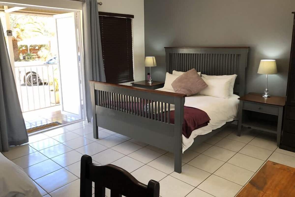 Un dormitorio con cama, dos mesitas de noche, una silla, ventana con persianas y vista al exterior.