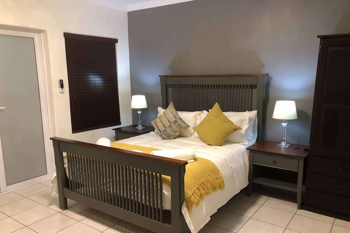 Un dormitorio con cama, dos mesitas de noche, una mesita auxiliar, cabecero de madera, ventana con persianas y una puerta.