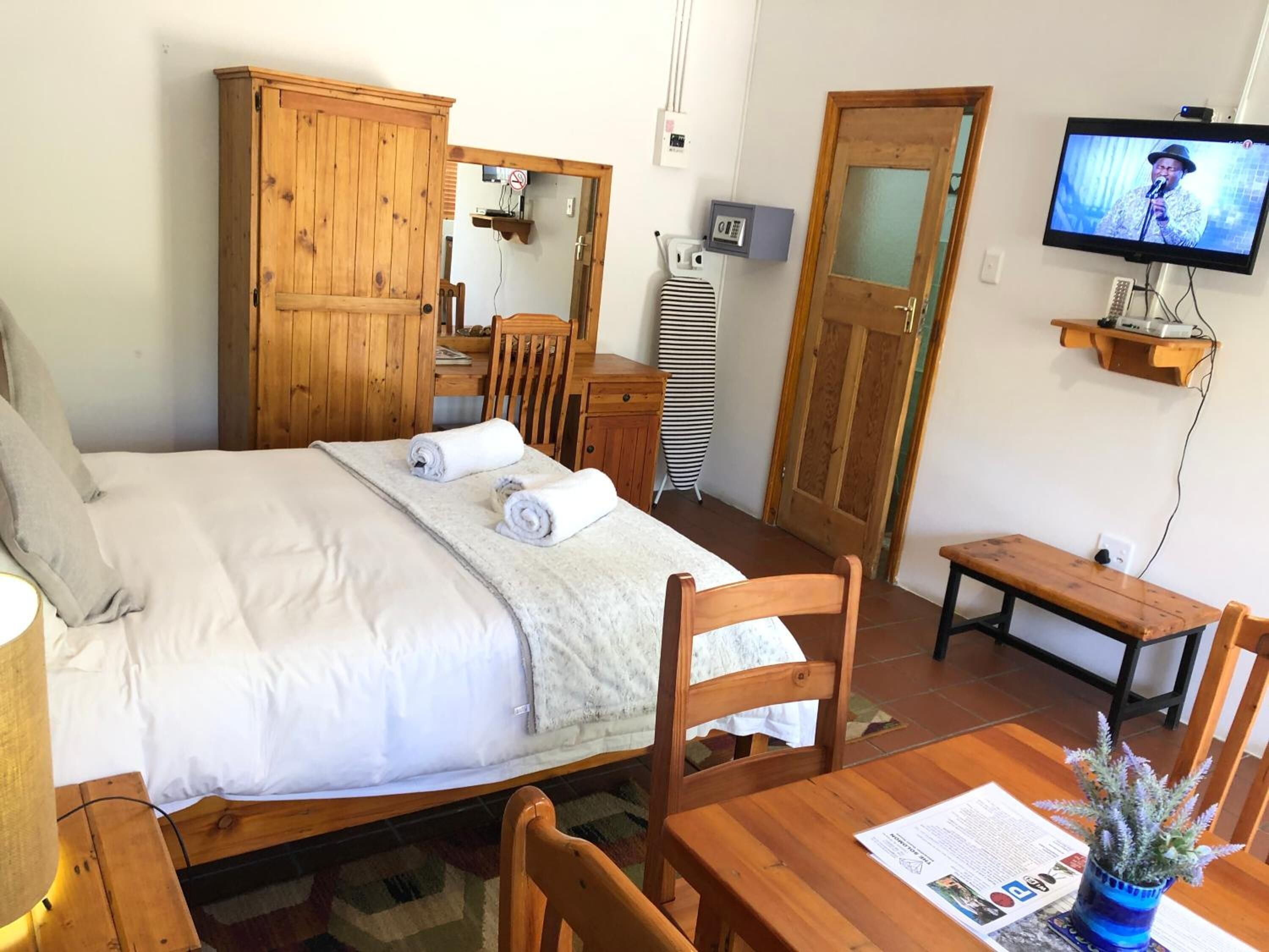 A bedroom with a bed, wooden wardrobe, a desk, a chair, a television, and a small table with a plant.